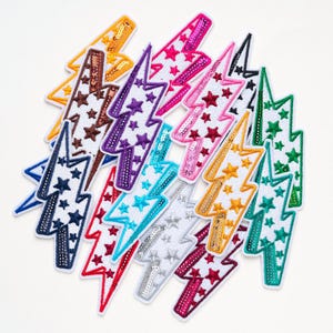 May include: A collection of lightning bolt-shaped patches in various colours including yellow, brown, purple, pink, black, blue, red, turquoise, silver, and green. Each patch features a white background with embroidered stars and sequin accents.