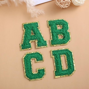 Può includere: Quattro toppe con lettere in tessuto verde: A, B, C e D. Ogni lettera ha un aspetto strutturato e sfocato con un bordo dorato glitterato. Le lettere sono disposte su una superficie beige chiaro, adatte per lavori manuali o personalizzazione.
