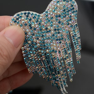May include: A heart-shaped embellishment covered in iridescent rhinestones, transitioning from clear to teal. Long, dangling fringe of teal rhinestones hangs from the bottom. The accessory is held up by a hand.