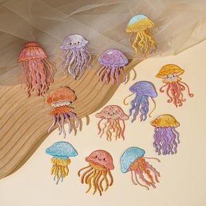 May include: A collection of eleven embroidered jellyfish patches in various colors including orange, purple, blue, and yellow. Each patch features a rounded bell top and dangling tentacles, with some designs displaying a small, simple smiling face.