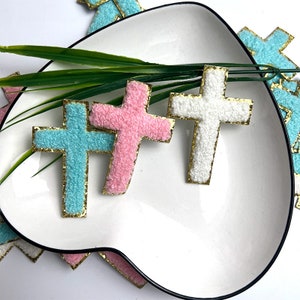 May include: Three felt cross shapes with gold trim. The crosses are pink, white, and light blue.