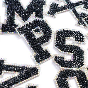 Puede incluir: Primer plano de varias letras y números negros de pedrería. Las letras están delineadas con pedrería iridiscente, creando un efecto brillante. Las letras están sobre un fondo blanco.