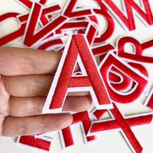 May include: Red felt letter patches with white outlines. The letter A is being held in a hand. The patches are ideal for adding a personalised touch to clothing, bags, or other crafts.