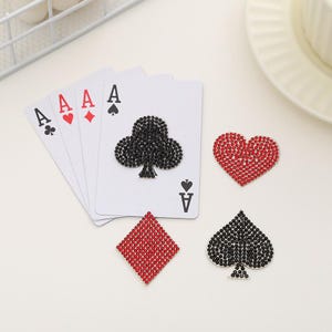 May include: Four playing cards with the letter A and a suit symbol, a black clover, a red heart, a red diamond, and a black spade.  There are also four rhinestone-covered playing card symbols: a black spade, a black clover, a red heart, and a red diamond.