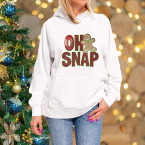 Op de afbeelding: Witte hoodie met een rood en groen ontwerp met de tekst "Oh Snap" en een gingerbread man-afbeelding.