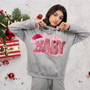 May include: A gray hoodie with the text "Santa BABY" in pink sequins, featuring a Santa hat. The hoodie has a front pocket and a drawstring hood. Christmas-themed apparel.