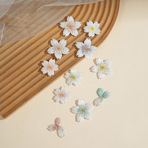 May include: A collection of white embroidered cherry blossom patches featuring five-petaled flowers with centers in various pastel colors including pink, yellow, purple, green, and blue, along with two small three-petal floral accent pieces.