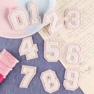 May include: Ten pink felt patches with gold embroidery, each featuring a number from 0 to 9. The patches are arranged in a random order on a blue denim background.
