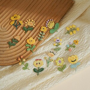 May include: A collection of small, colorful embroidered flower patches featuring various yellow, orange, and purple blossoms with green stems and leaves. Each patch displays a unique floral design, including sunflowers, daisies, and buds.