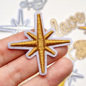 May include: Embroidered star patch with gold thread on a light blue background. The star has eight points and a white border. Other patches with the word "love" and an anchor are visible in the background.