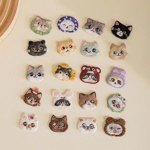 May include: A collection of twenty embroidered cat face patches in various colors and designs, including cats wearing animal hoods, hats, and glasses. Each patch features detailed stitching of feline facial features like eyes, noses, and whiskers.