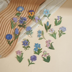 Può includere: Una collezione di ventuno toppe ricamate a forma di fiore con vari disegni botanici in tonalità di blu, viola, rosa e giallo, ciascuna con steli e foglie verdi, disposte in modo sparso su una superficie di colore chiaro.