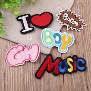 May include: Six embroidered iron-on patches with various designs. The patches include a red heart with the word "I" in white, the word "Boy" in green, yellow, and white, the word "Girl" in pink, the word "Music" in red, blue, and yellow, and the word "Boom" in brown.
