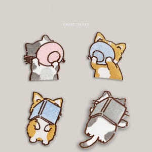 May include: Four embroidered cat patches in different poses, each holding an object. The cats are in shades of grey, orange, and white, with the objects in pink, blue, and grey. The word "EMBROIDERED" is above the patches.