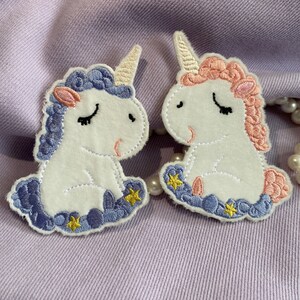 May include: Two embroidered unicorn patches, one with blue and purple accents and the other with pink accents. Both unicorns are sitting with their eyes closed and have gold stars around them.