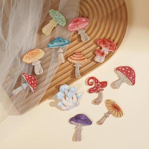 May include: A collection of twelve embroidered mushroom patches in various colors including red, blue, green, orange, and purple. Each patch features detailed stitching, depicting different mushroom shapes, caps, and stems in a variety of sizes.