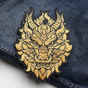 May include: Embroidered patch featuring a golden dragon head with intricate details. The dragon has horns, a detailed mane, and a fierce expression. The patch is set against a dark blue denim background.