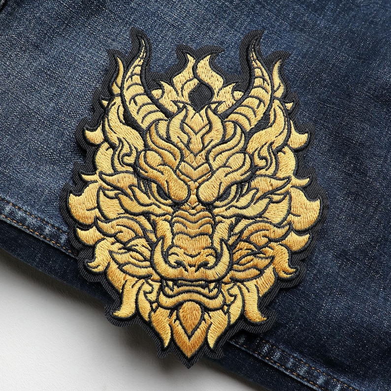 May include: Embroidered patch featuring a golden dragon head with intricate details. The dragon has horns, a detailed mane, and a fierce expression. The patch is set against a dark blue denim background.