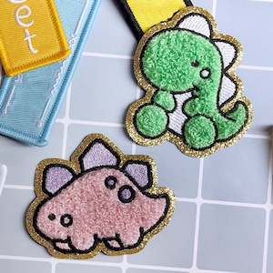 Könnte beinhalten: Zwei Plüsch-Dinosaurier-Patches mit goldfarbenem Glitzerbesatz. Einer ist grün und der andere ist rosa mit violetten Akzenten.