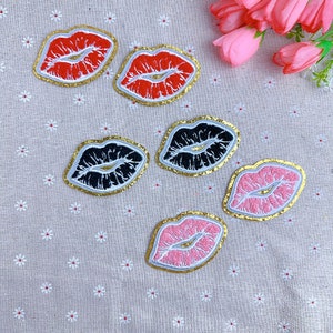 May include: Embroidered lip patches in red, black, and pink with gold trim. The patches are in various sizes and arranged on a light beige fabric with small white flower patterns. A bouquet of pink flowers is in the background.