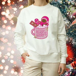 Pode incluir: Camisola branca com um design bordado rosa de uma caneca com chocolate quente, marshmallows, um bastão de caramelo e um homem de gengibre. A caneca diz "Merry & Bright".
