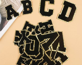 Puede incluir: Un conjunto de parches de letras de fieltro negro con ribete de purpurina dorada. Las letras están en una fuente de estilo universitario. Las letras incluyen A, B, C, D, E, F, G, H, I, J, K, L, M, N, O, P, Q, R, S, T, U, V, W, X, Y y Z.