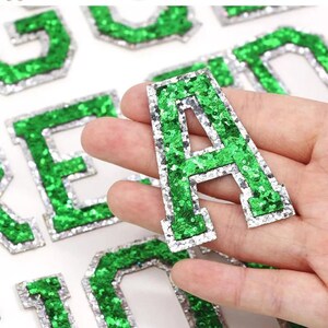 May include: Green and silver glitter iron-on letters. The letters are in a varsity style font. The letters are perfect for adding a personal touch to clothing, bags, and other accessories.