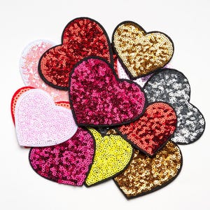 May include: A collection of heart-shaped sequin patches in various colors, including red, pink, gold, and silver. Each patch has a black border, creating a defined shape. The sequins catch the light, giving a sparkling effect. These patches are suitable for crafts and embellishments.