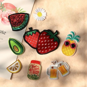May include: A selection of embroidered patches with diverse designs. These include a watermelon slice with green sequins, two strawberries, an avocado, a lemon slice, a daisy, a pineapple with sunglasses, a jar with text, and two beer mugs.