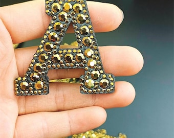 Può includere: Un'applicazione nera a forma di lettera "A", fittamente ricoperta di strass color oro. La lettera è tenuta in mano, mostrando i dettagli scintillanti degli abbellimenti. Questa applicazione decorativa è adatta a vari progetti artigianali.