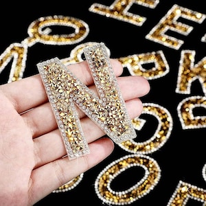 May include: A close-up of a hand holding a gold letter N made of rhinestones. The letter is surrounded by other letters made of rhinestones, including E, F, and K.