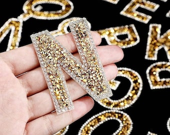 Può includere: Primo piano di una mano che tiene una lettera N dorata fatta di strass. La lettera è circondata da altre lettere fatte di strass, tra cui E, F e K.