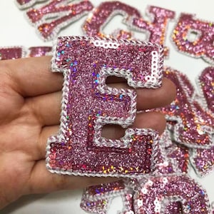 May include: A pink glittery fabric letter "E" with a white sequined border and white stitching. The letter is held in a hand, with other letters visible in the background. The letters are likely for crafts or personalization.