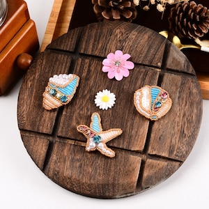 May include: Assortment of five beaded embellishments in the shape of a seashell, starfish, and two flowers. The embellishments are on a wooden surface.