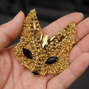 May include: A gold fox-shaped applique with black rhinestone eyes. The applique is covered in small gold rhinestones.