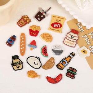 Op de afbeelding: Een verzameling kleine geborduurde patches met diverse etenswaren, waaronder popcorn, een drankje, een zak chips met het label WA, een melkfles, een stokbrood, een watermeloenpartje, een gebakken ei, een burger, een worstje en andere snacks.