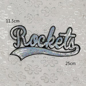 Op de afbeelding: Een zilveren pailletten-strijk-op-patch met het woord "Rockets" in een zwarte omtrek. De patch meet 11,5 cm bij 25 cm.