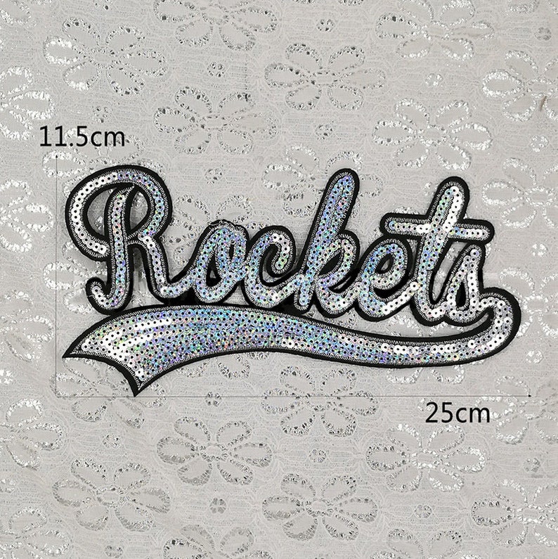 Op de afbeelding: Een zilveren pailletten-strijk-op-patch met het woord "Rockets" in een zwarte omtrek. De patch meet 11,5 cm bij 25 cm.