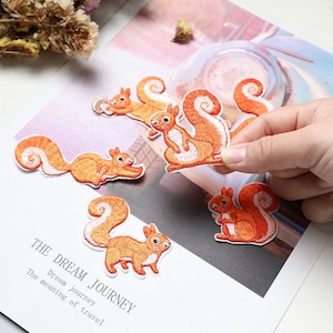 May include: Embroidered patches featuring orange squirrels with white accents. The patches have detailed stitching and various poses. The text "THE DREAM JOURNEY" is visible on a white background.
