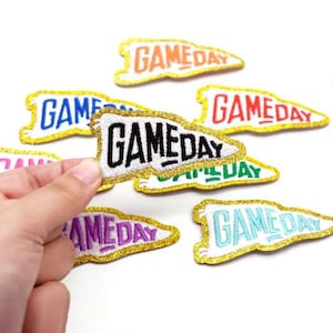 Könnte beinhalten: Ein Set mit sieben Bügelbildern in verschiedenen Farben mit einem goldenen Glitzer-Umrandung. Jedes Bügelbild hat die Form eines Wimpels mit dem Text "GAMEDAY" in verschiedenen Farben und Schriftarten.