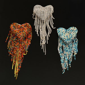 May include: Three rhinestone heart-shaped brooches with cascading fringe. One brooch is silver, another is a mix of red, orange, yellow, and blue, and the third is blue and white. All three are set against a black background.