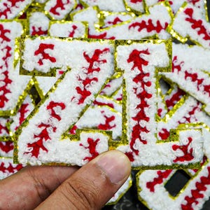 Op de afbeelding: Close-up van een wit, rood en goud geborduurde letter "Z" patch. De letter is gevuld met wit, pluizig materiaal en rode stiksels, omlijnd met gouden glitter. De patch wordt door een hand vastgehouden, met andere soortgelijke patches op de achtergrond.