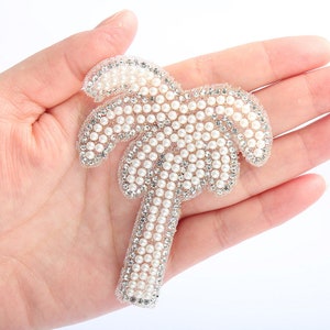 May include: A white palm tree-shaped patch made of pearls and rhinestones. The patch is on a hand, but the focus is on the patch itself.