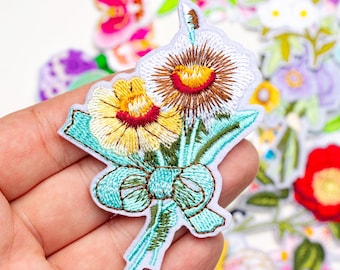 May include: Embroidered floral patch featuring a bouquet of flowers with yellow, brown, and red centers, tied with a turquoise bow. The patch is white with detailed stitching and is surrounded by other floral patches.