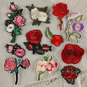 May include: Assortment of embroidered iron-on patches featuring various floral designs. The patches include red, pink, white, and purple flowers with green leaves.