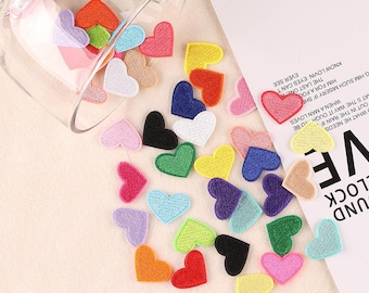 May include: A collection of colorful felt heart patches in various shades of red, pink, blue, green, yellow, and purple. The patches are arranged in a glass jar and on a white surface.