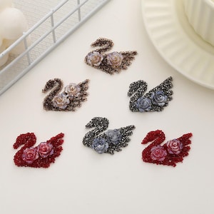 May include: Six swan-shaped embellishments with rhinestone accents and faux pearl flower centres. The swans are in various colours including brown, grey, blue, and red.
