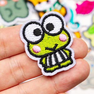 May include: Embroidered patch featuring a cartoon frog with large eyes, pink cheeks, and a striped belly. The patch has a white border and is primarily green, black, and white. Other colorful patches are visible in the background.