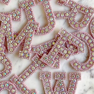 May include: Pink, rhinestone-covered letters scattered across a white surface. The letters are in various fonts and sizes, with each letter individually adorned with sparkling rhinestones. The letters are a light pink color.