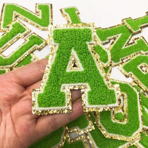 Op de afbeelding: Groene vilt letter patches met gouden glitterrand. De letter 'A' wordt in een hand gehouden.